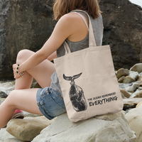 Person sitting on rocks by the ocean holding a tote bag with a whale design and text.